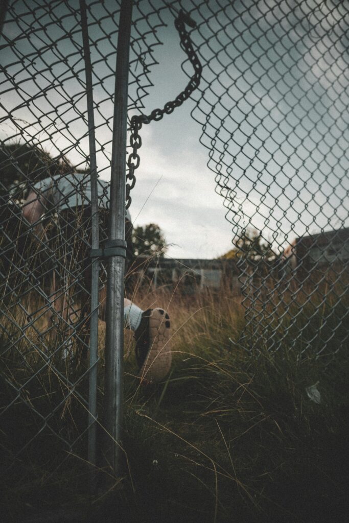 A person exploring an abandoned area through a broken chain link fence.